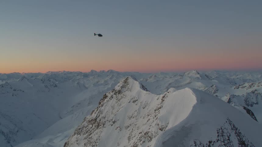 Helicopter flies over snow covered mountain range against a red tinted sky