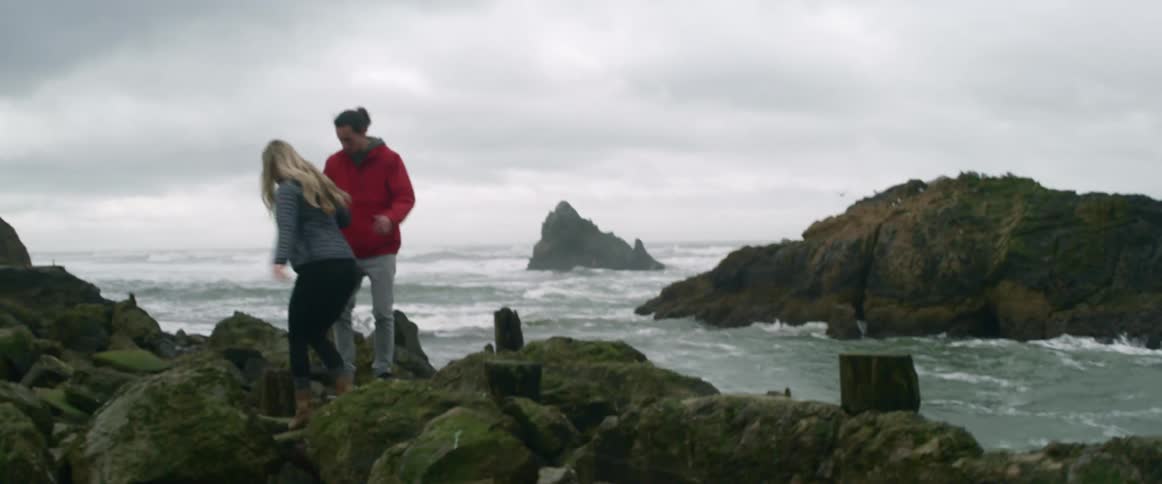 A man helps a woman across some rocks by the shore
