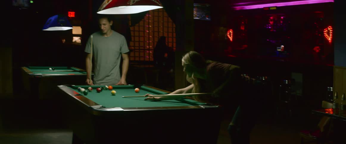 A man cheers a woman on as she shoots pool in a bar