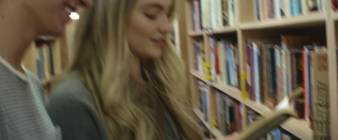 A man and woman stand in a library reading a book and talking