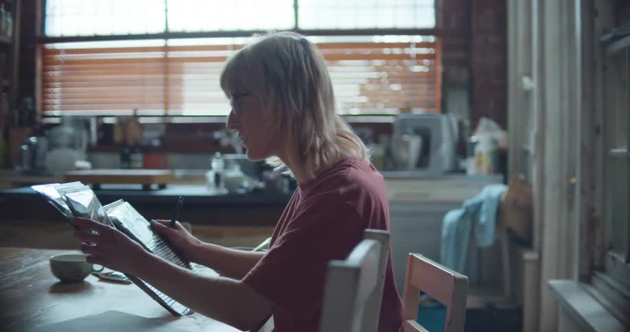 A woman sits at her kitchen table and goes through a photo album while writing information down on paper