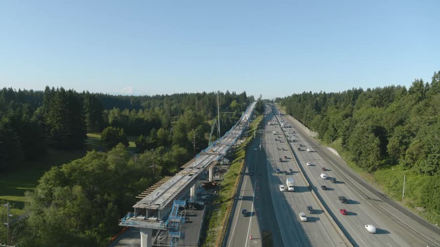 Vehicles drive on a highway that parallels an elevated light rail deck under construction through a green suburban neighborhood