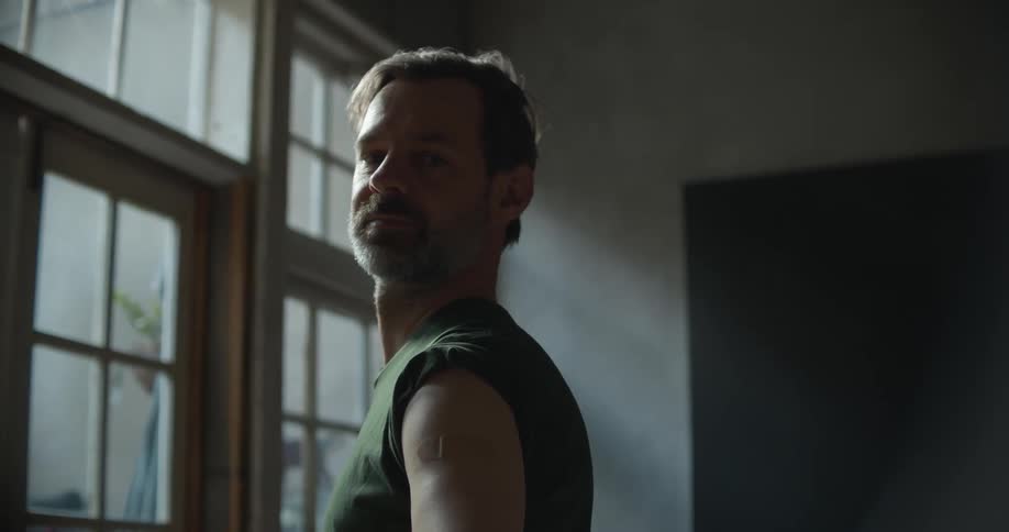 A man stands indoors with his shirt sleeve rolled up and a bandage on his arm