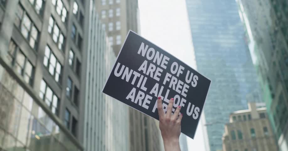 Protester with red fingernail polish holds sign up standing between downtown skyscrapers