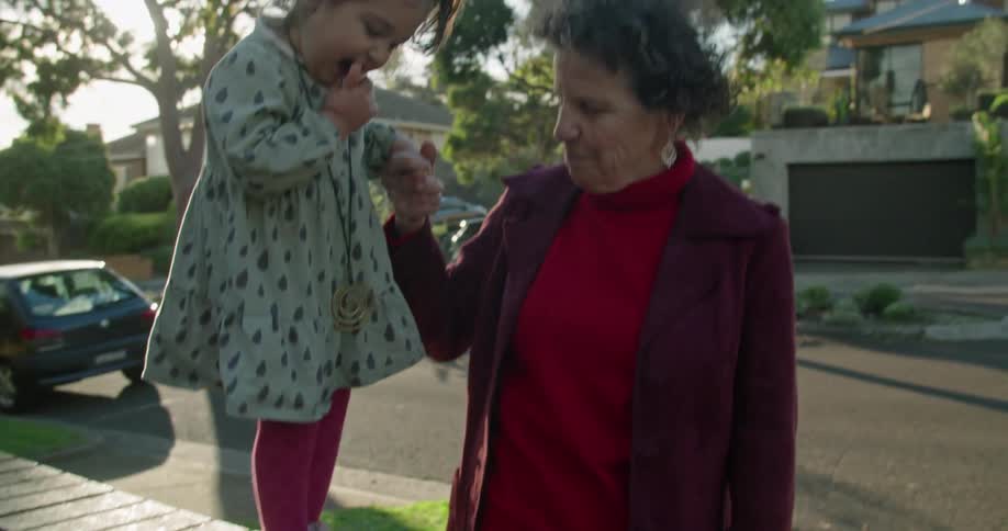A woman holds her granddaughters hand as the toddler walks along a ledge in their yard