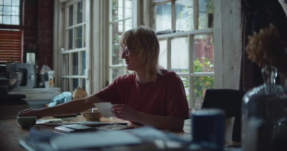 A woman sits at a desk in a home drinking from a cup and looking at papers