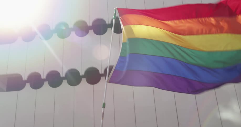 People march carrying rainbow flag past building decorated with series of circular opening on bright day