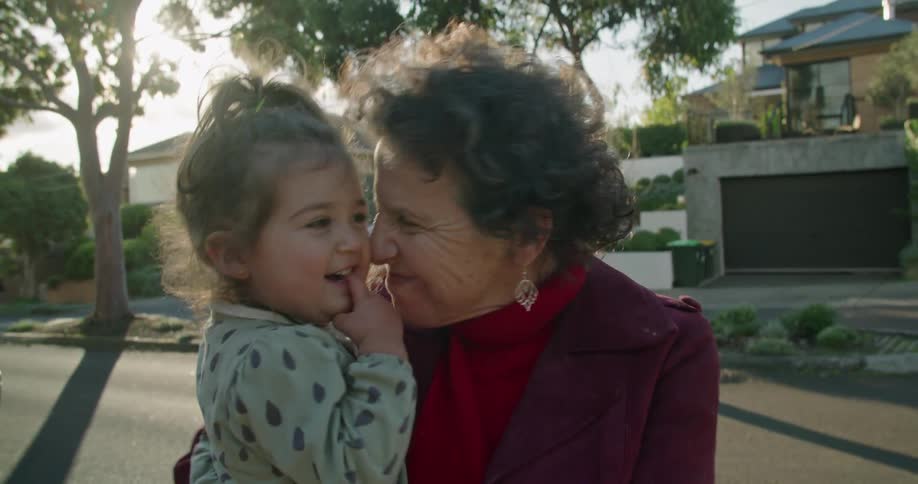 A grandmother smiles and sways while holding her granddaughter on a sunny sidewalk in a neighborhood
