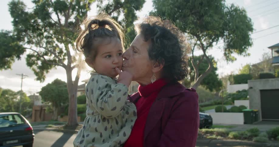 A woman holding and kissing a little girl with trees houses the sky and clouds in the background
