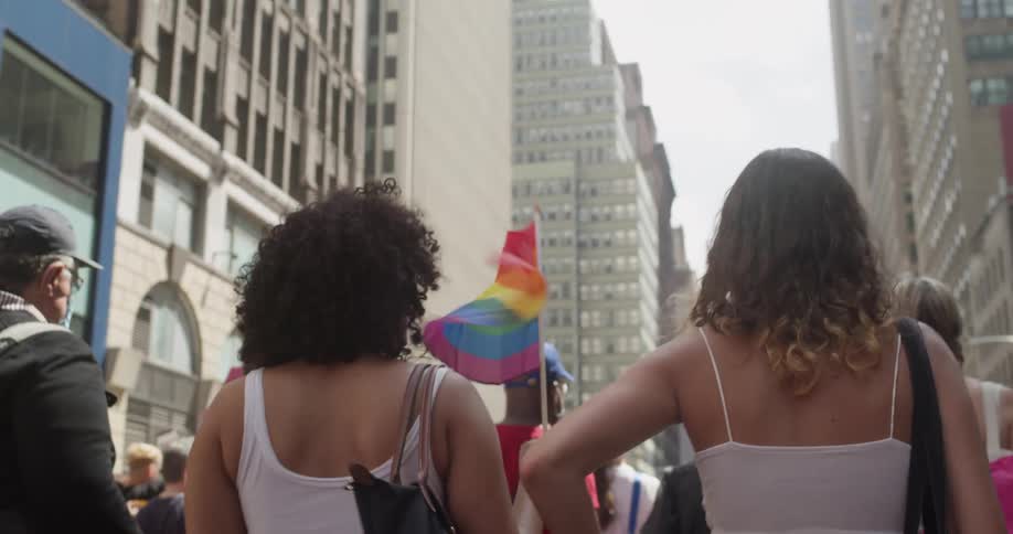 Two women carrying flag and taking pictures walk among crowd through downtown at Pride event