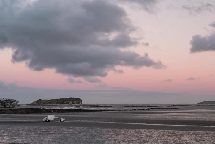 A boat sits on the sand as waves come to shore and clouds pass overhead