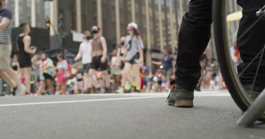 A person waits with a bicycle beside a road as a pride parade walks by