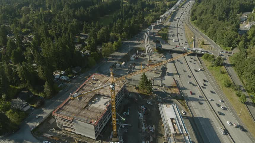 Highway traffic flows past a new light rail station under construction near a tree lined suburban neighborhood with a city skyline on the horizon