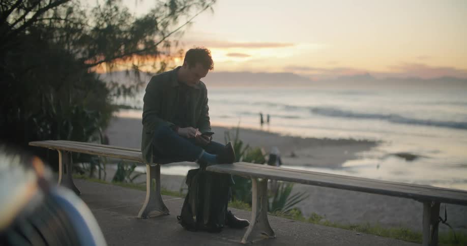 A man typing on a smartphone while sitting on a bench near the sea
