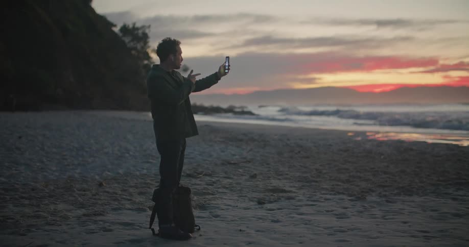 A man waves during a video call as he stands on the beach