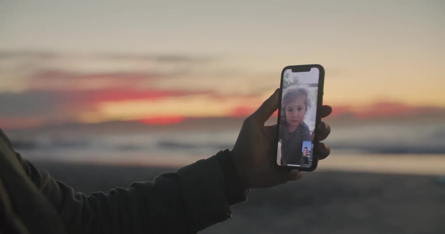 Man stands on beach talking and waving at girl on video call on phone with red sky on horizon