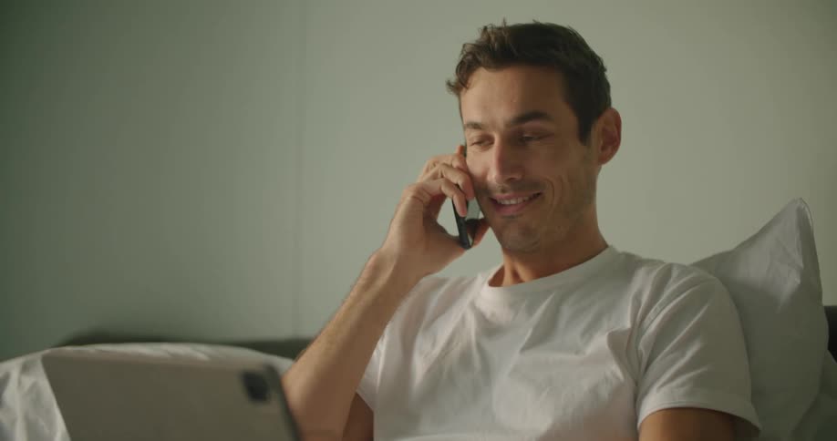 A man smiles while sitting up in bed talking on the phone and working on a laptop