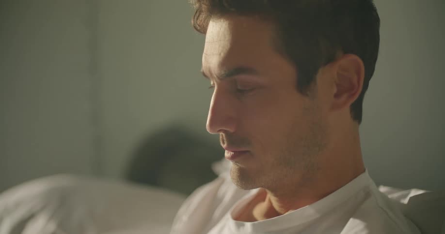 A man sighs and nods a bit while typing on his laptop in bed