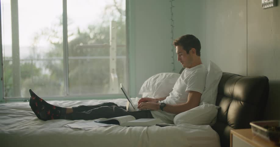 A man types on his laptop in bed with a book and notes spread out around him
