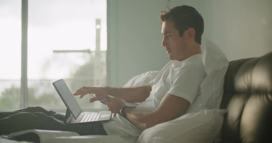 A man takes notes while sitting up in bed working on his laptop