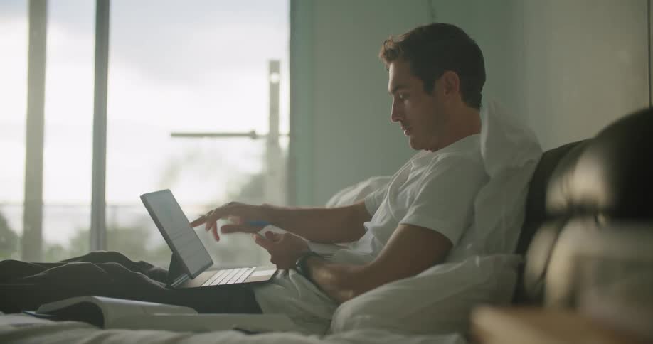 A man writes down information from his laptop while lying in bed