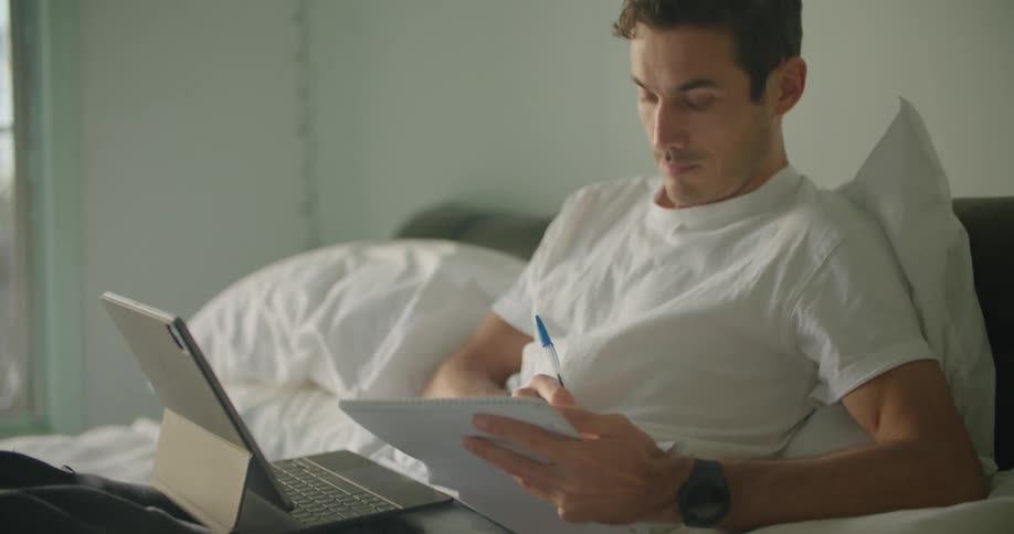 A man reclines in bed with his laptop and books while he takes notes