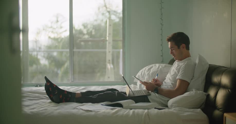 A man sits up in a bed writing in a notebook while holding a laptop