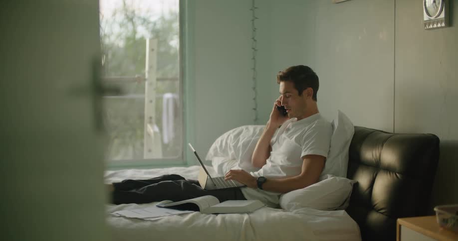 A man using a laptop while sitting on a bed answers his smartphone