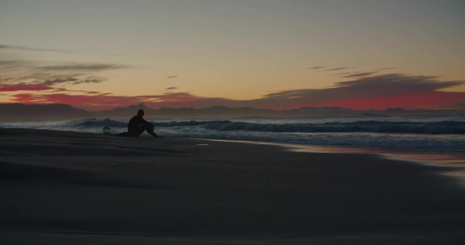 A surfer sitting on a sandy beach looks out over the waves contemplatively at sunset