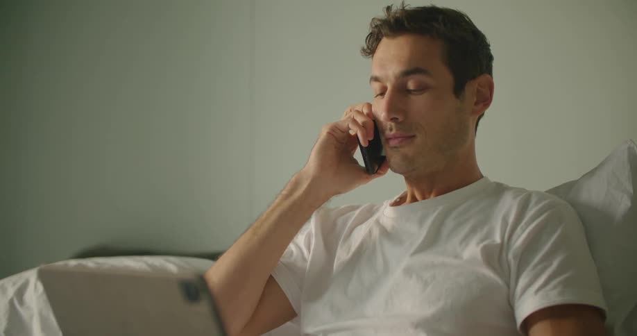 Man sits in bed and talks on his cell phone then puts the phone down and works on a laptop