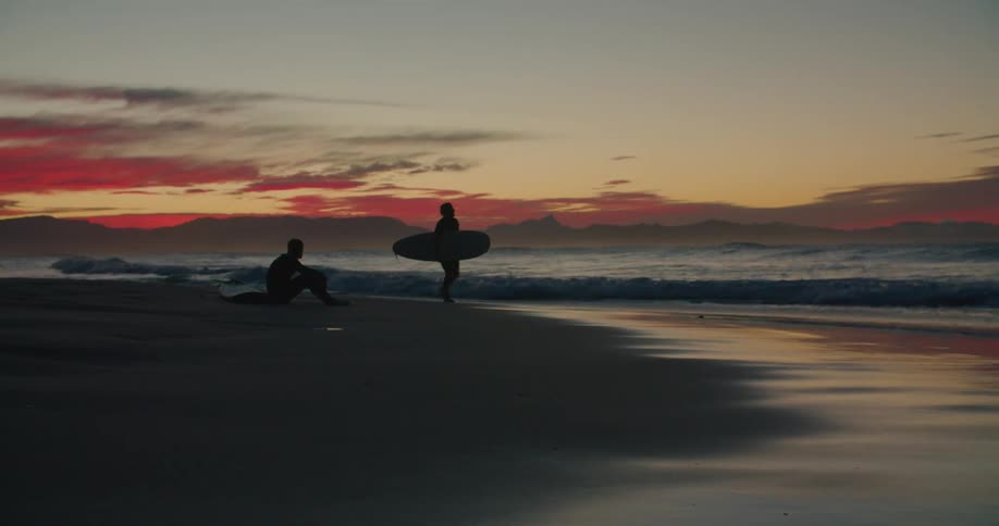 Crimson clouds hover over mountains on horizon as silhouette of surfer carrying surfboard walks past male silhouette sitting on the beach
