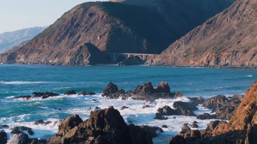 Aerial view of waves crashing against rocks at the base of mountains