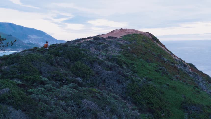 Blonde woman wearing red backpack walks to the top of the hill on a cloudy day