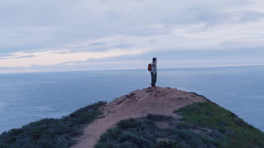 A woman wearing a hat stands on an outcropping of rock overlooking the ocean