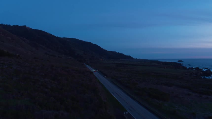 One lone car drives down the coast with the ocean to its right and beautiful rolling green hills on the left at sunrise