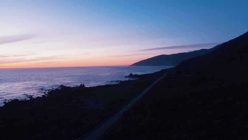 Single straight road runs along a mountainous coastline next to the ocean as the sun below the horizon casts beautiful colors across the sky