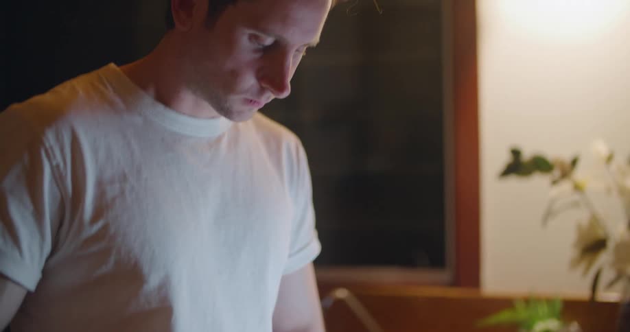 A man dices vegetables on a cutting board in the kitchen with his laptop open on the counter