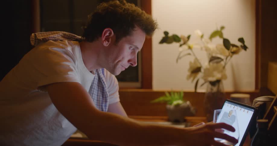 Man sits near dark window drinking wine and scrolling on computer