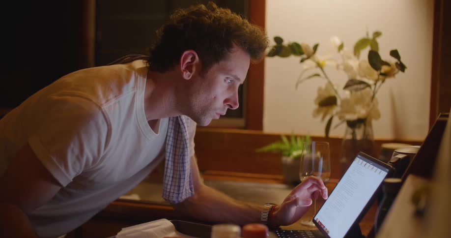 A man in the room looking at a laptop with a wall and window in the background