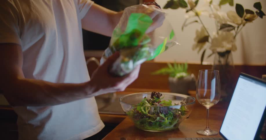 Man places greenery from plastic bag into salad bowl sitting near wine glass and laptop