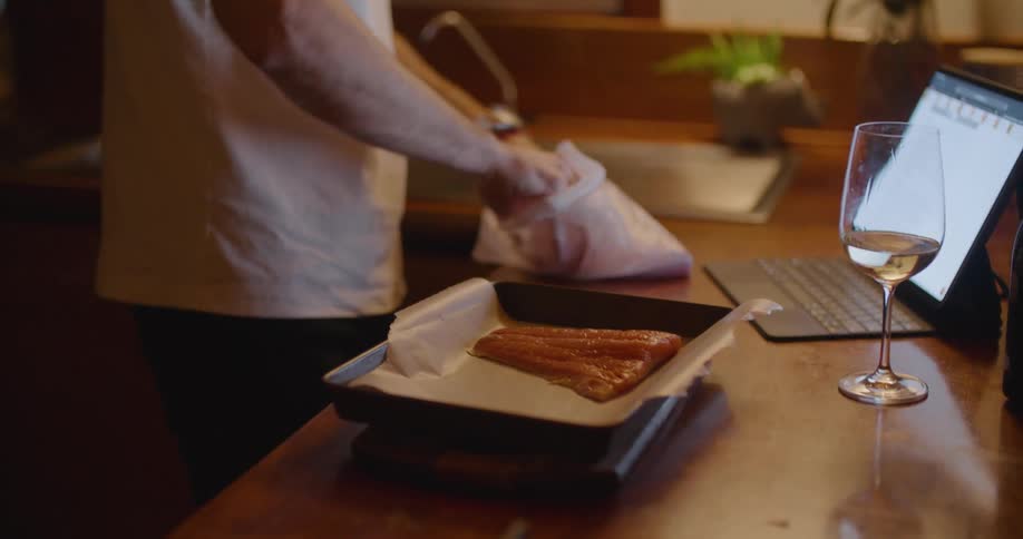 A man puts pieces of raw fish on a pan and looks at a laptop while taking a drink of wine in a home kitchen