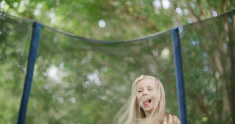 Blonde girl wearing pink dress jumping on a trampoline in the backyard