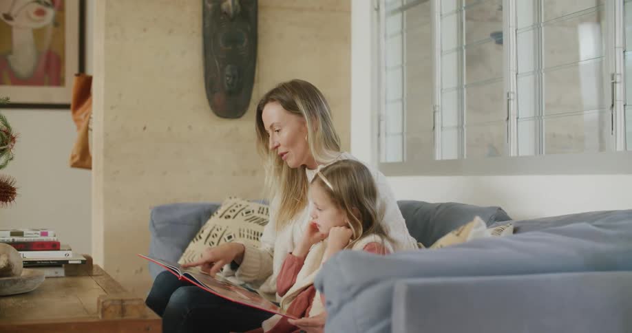 Daughter fiddles with hair as they sit on couch in living room and mom read from book