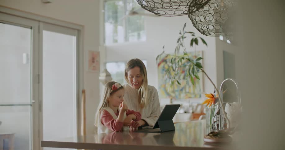 A woman uses a tablet with her daughter as they sit at their kitchen island