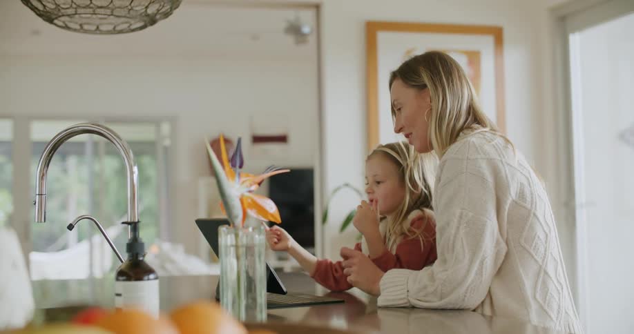 A woman and her daughter use a tablet on a stand on a counter in a home kitchen during the day