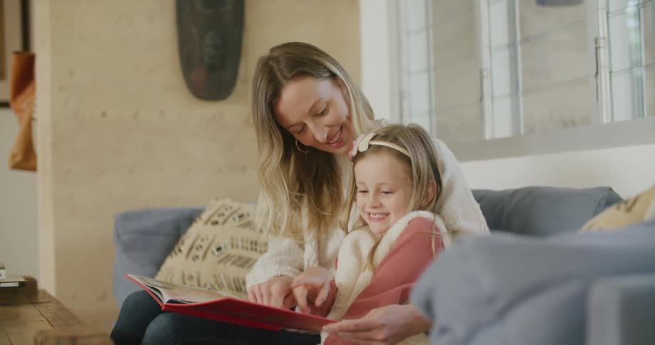 Woman wearing sweater hugs daughter as they sit on sofa sharing book