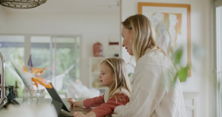 Blonde mother with white sweater helps daughter use tablet sitting at kitchen island at home