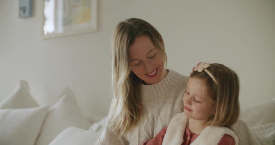 A mother sits on a couch at home with her daughter and smiles as her daughter uses a tablet