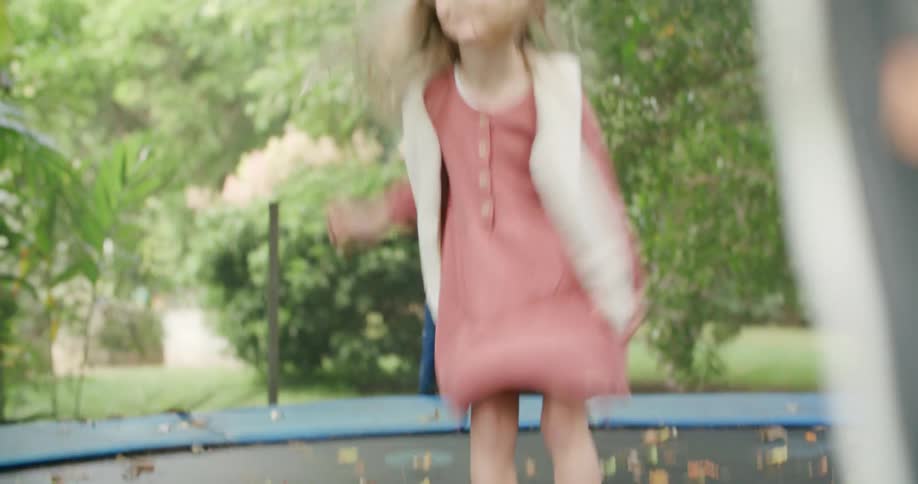 Blonde girl wearing vest and dress bounces on trampoline surrounded by protective netting and strewn with fallen leaves