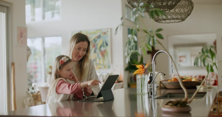 Mother and daughter sit at kitchen island across from sink using a tablet